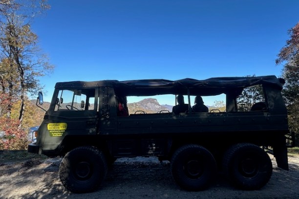 Military-style 6x6 vehicle with passengers, parked on dirt road with trees and mountains in background.
