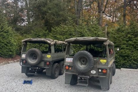 Two parked camouflage military vehicles on gravel surrounded by trees.