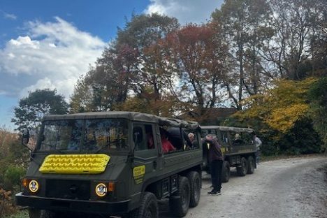 Two military-style vehicles on a dirt road lined with autumn trees under a partly cloudy sky.